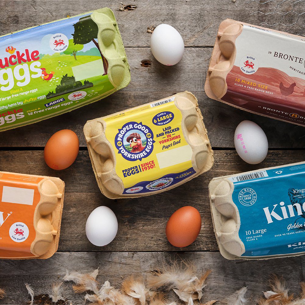 A range of egg packs from several brands laid out on a wooden surface with loose eggs and feathers.